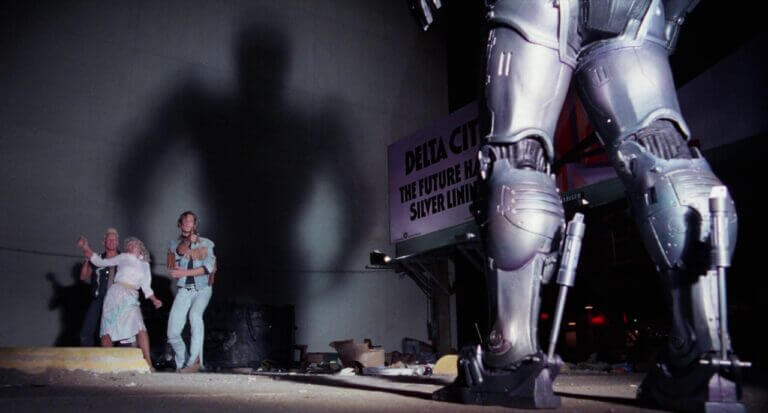 RoboCop stands before and his shadow towers above two criminals who are harassing a woman