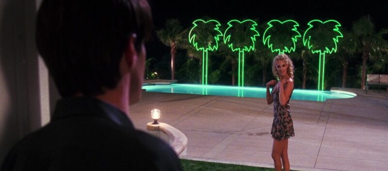 Nomi looks over at Zack while standing outside a pool lit up by neon palm trees