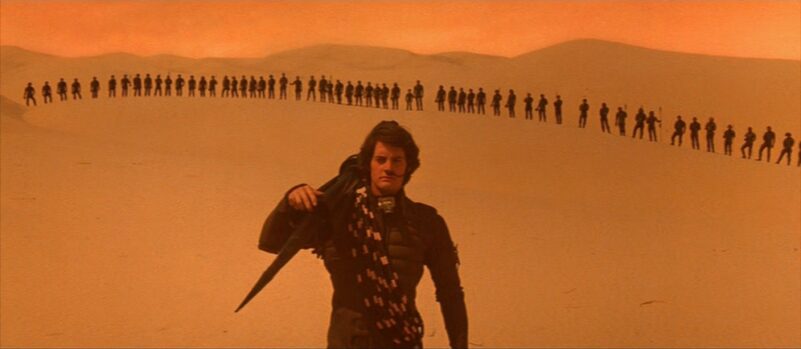 Paul Atreides walks through a desert as several people stand in the background in Dune