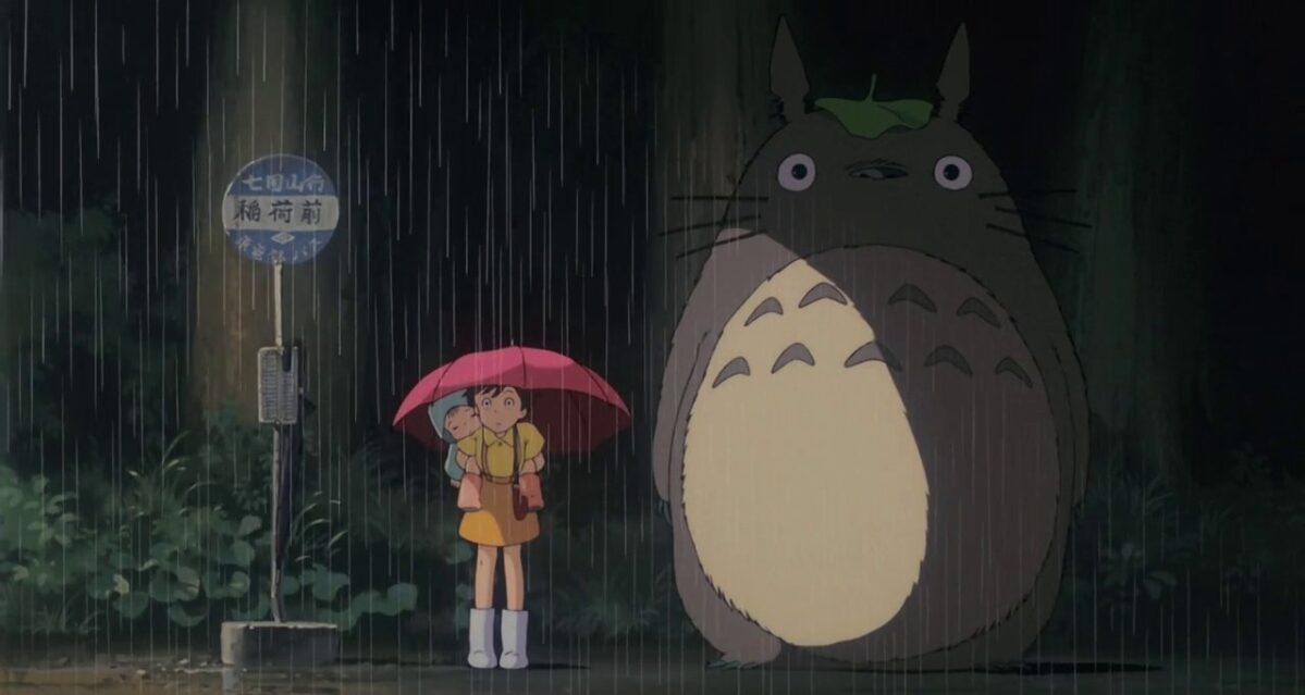 Satsuki stands holding an umbrella while carrying her sister May next to Totoro