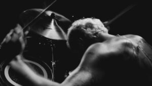 A shirtless drummer plays heavy metal