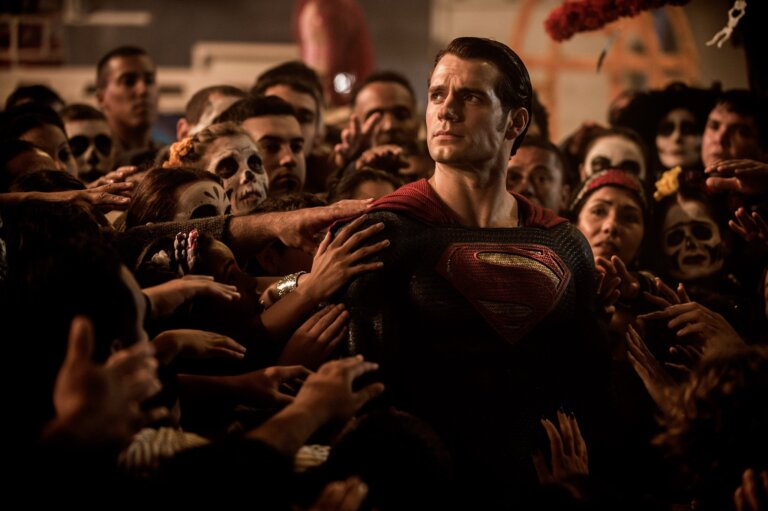 Superman stands in a crowd of people who all put a hand on him, in awe.